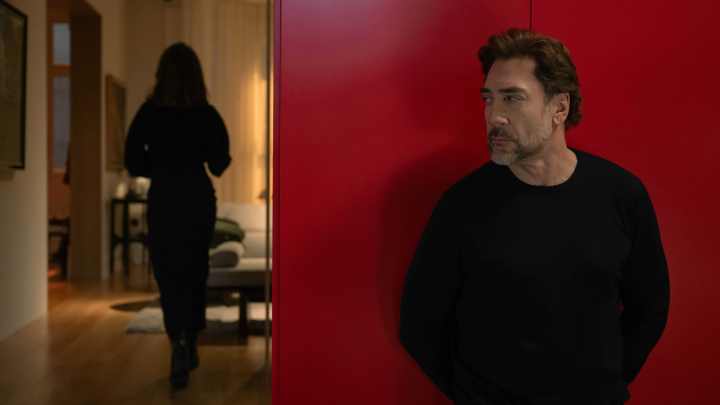 Ein Mann in schwarzem Pullover steht nahe einer roten Wand, während eine Frau in schwarzer Kleidung im Hintergrund geht.