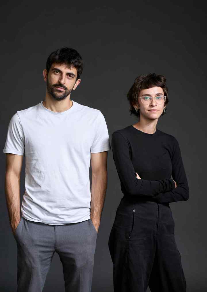 Ein Mann und eine Frau stehen nebeneinander vor einem dunklen Hintergrund. Der Mann trägt ein weißes T-Shirt und graue Hose, während die Frau ein schwarzes Oberteil und schwarze Hose trägt. Beide schauen neutral in die Kamera.