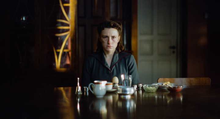 Eine Frau sitzt an einem Tisch, der mit verschiedenen Lebensmitteln und Getränken gedeckt ist. Sie schaut ernst und nachdenklich in die Kamera. Der Hintergrund ist dunkel und vermittelt eine melancholische Stimmung.