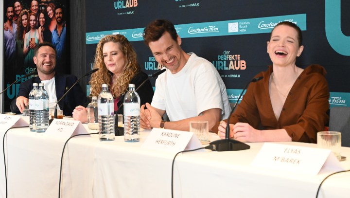 Pressekonferenz für den Film 'Der perfekte Urlaub' mit mehreren Schauspielern, die an einem Tisch sitzen und miteinander lachen.
