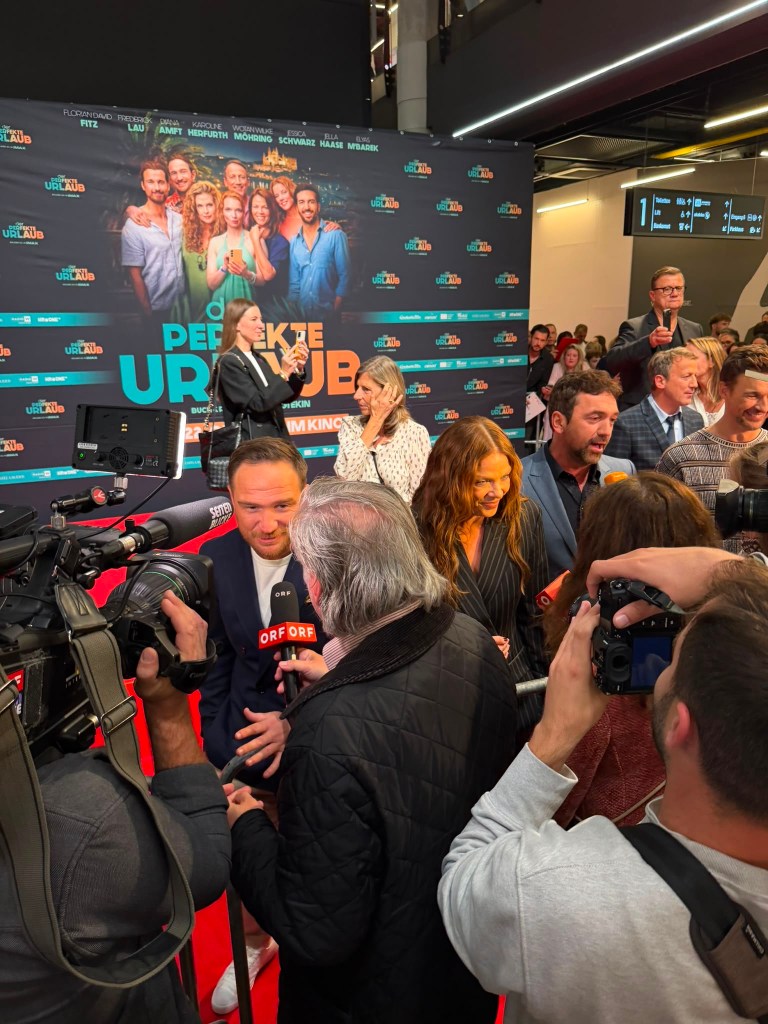 Premierenveranstaltung mit einem roten Teppich, auf dem ein Mann ein Interview gibt, während Kameraleute und Publikum zuschauen. Im Hintergrund eine große Wand mit dem Titel 'Perfekte Urlaub' und den Namen der Darsteller.